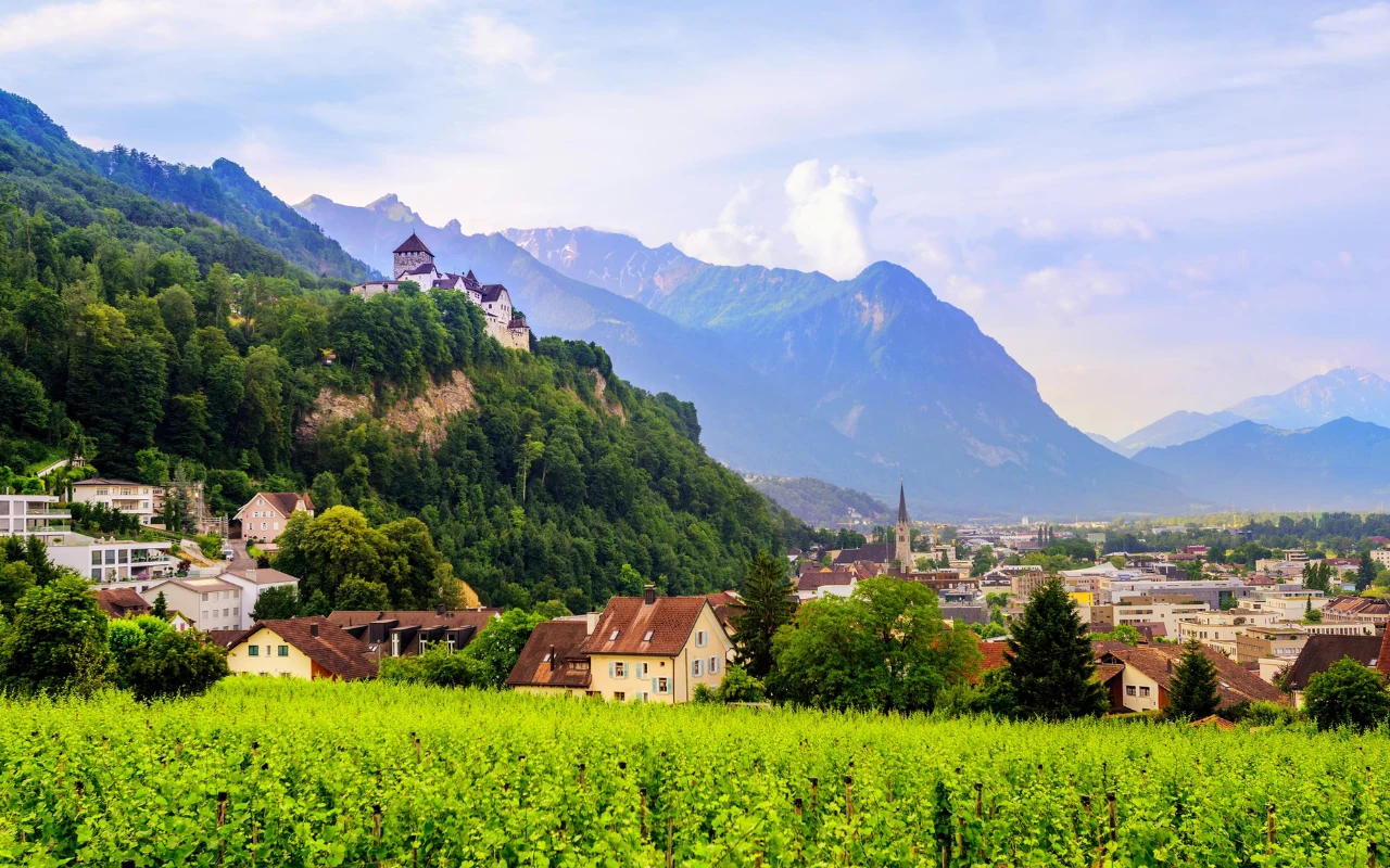 Vaduz Zamek w Vaduz, położony na wzgórzu, wśród bujnej zieleni, poniżej rozległa zabudowa miasta, w tle alpejskie szczyty.