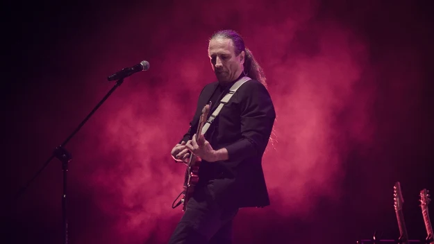 Gitarzysta z długimi włosami gra na elektrycznej gitarze na scenie, otoczony purpurowym światłem i dymem, obok niego ustawiony mikrofon oraz dwa stojące gitary w tle.