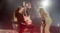 Gitarzysta w dynamicznej pozie gra na czerwonej gitarze elektrycznej, obok wokalistka pochylona z mikrofonem, scena oświetlona jasnym światłem reflektorów.