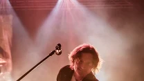 Gitarzysta w dynamicznej pozie podczas koncertu, trzymający instrument i wyciągający nogę do tyłu, podświetlony intensywnym światłem scenicznym w odcieniach różu i żółci.