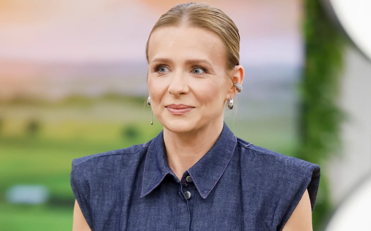 Kobieta z blond włosami upiętymi do tyłu, ubrana w ciemnoniebieską, bezrękawną koszulę na tle rozmytej, zielonej scenerii.