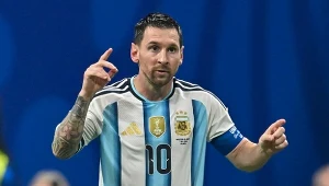 Piłkarz w biało-błękitnej koszulce Argentyny z numerem 10 unosi dłonie, patrząc przed siebie, z opaską kapitana na lewym ramieniu.