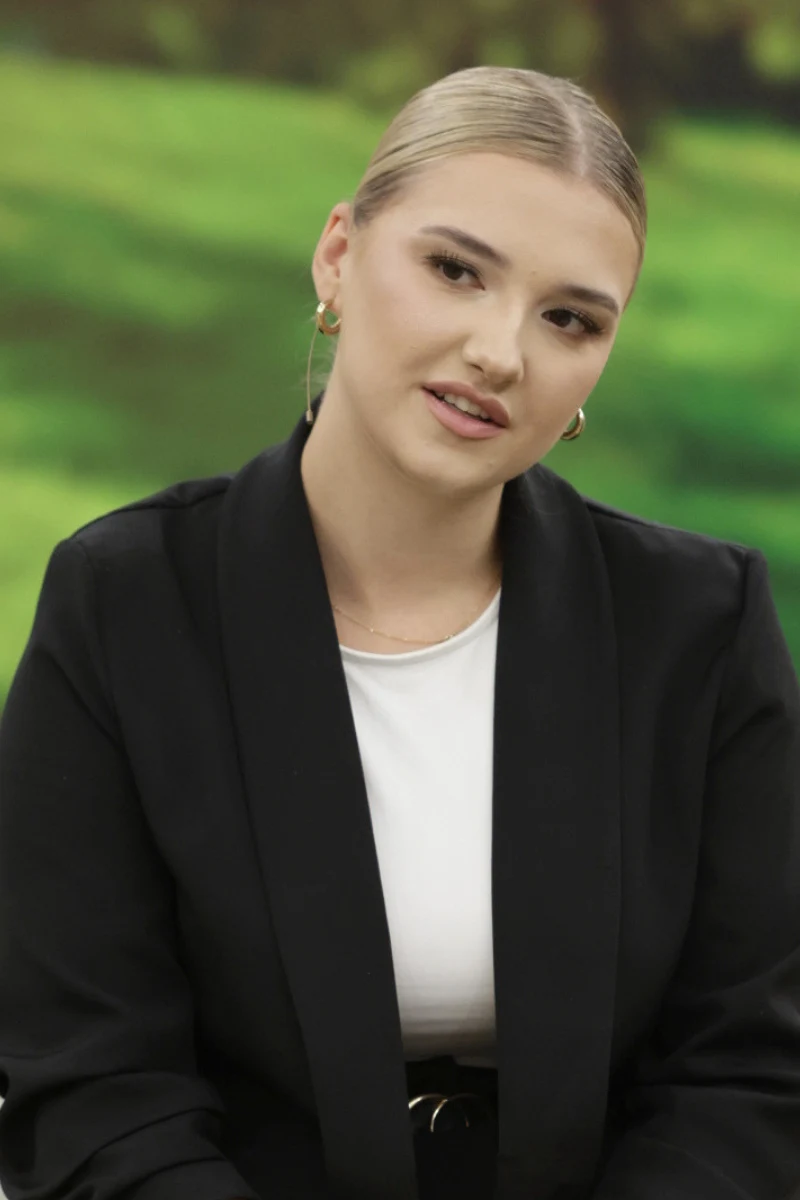 Młoda kobieta z jasnymi włosami spiętymi w gładki kok, ubrana w biały top i czarną marynarkę, patrzy lekko w bok z delikatnym uśmiechem na twarzy.
