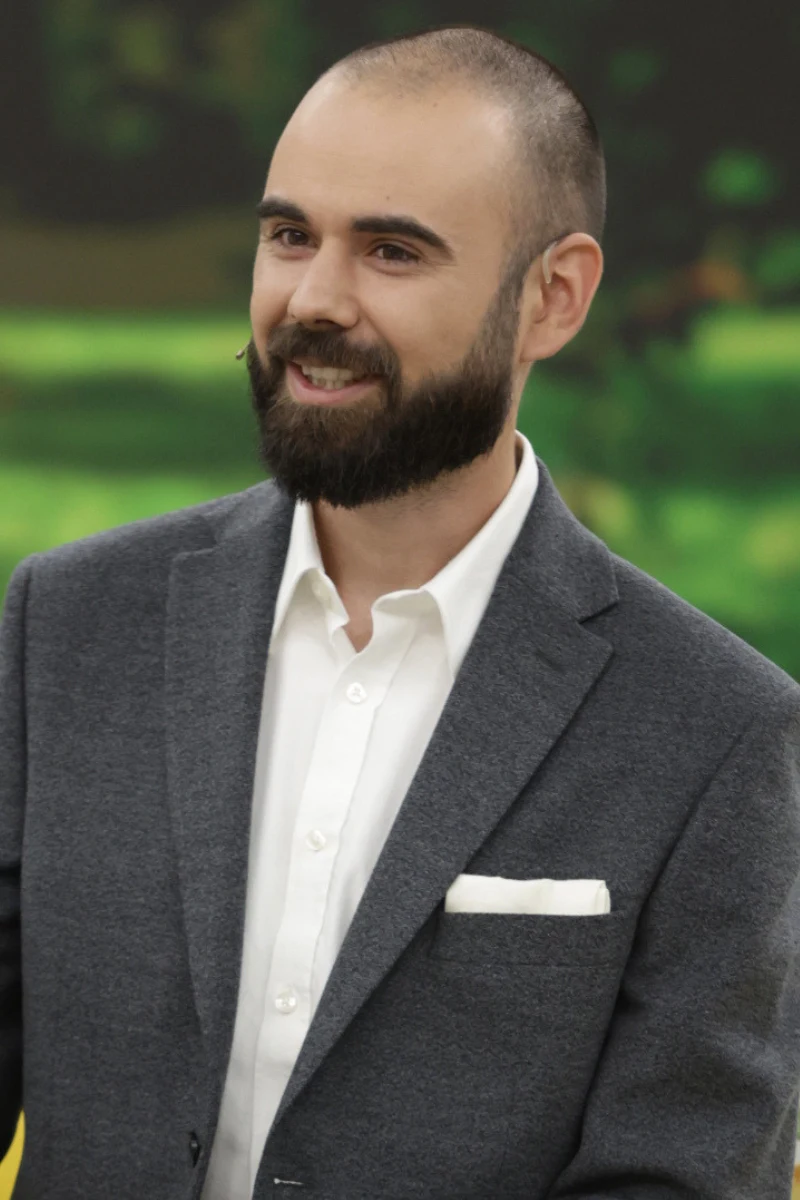 Uśmiechnięty mężczyzna z brodą w eleganckiej, szarej marynarce i białej koszuli, z białą poszetką w kieszeni marynarki.