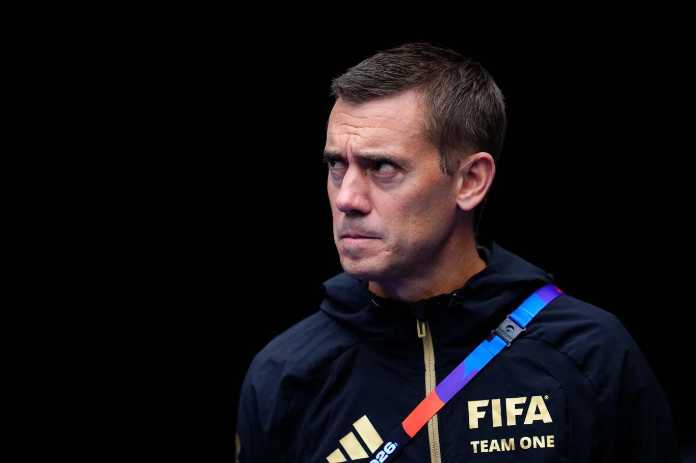 Mężczyzna w granatowej sportowej kurtce z napisem FIFA TEAM ONE na piersi, patrzący poważnym wzrokiem w bok, na ciemnym tle.