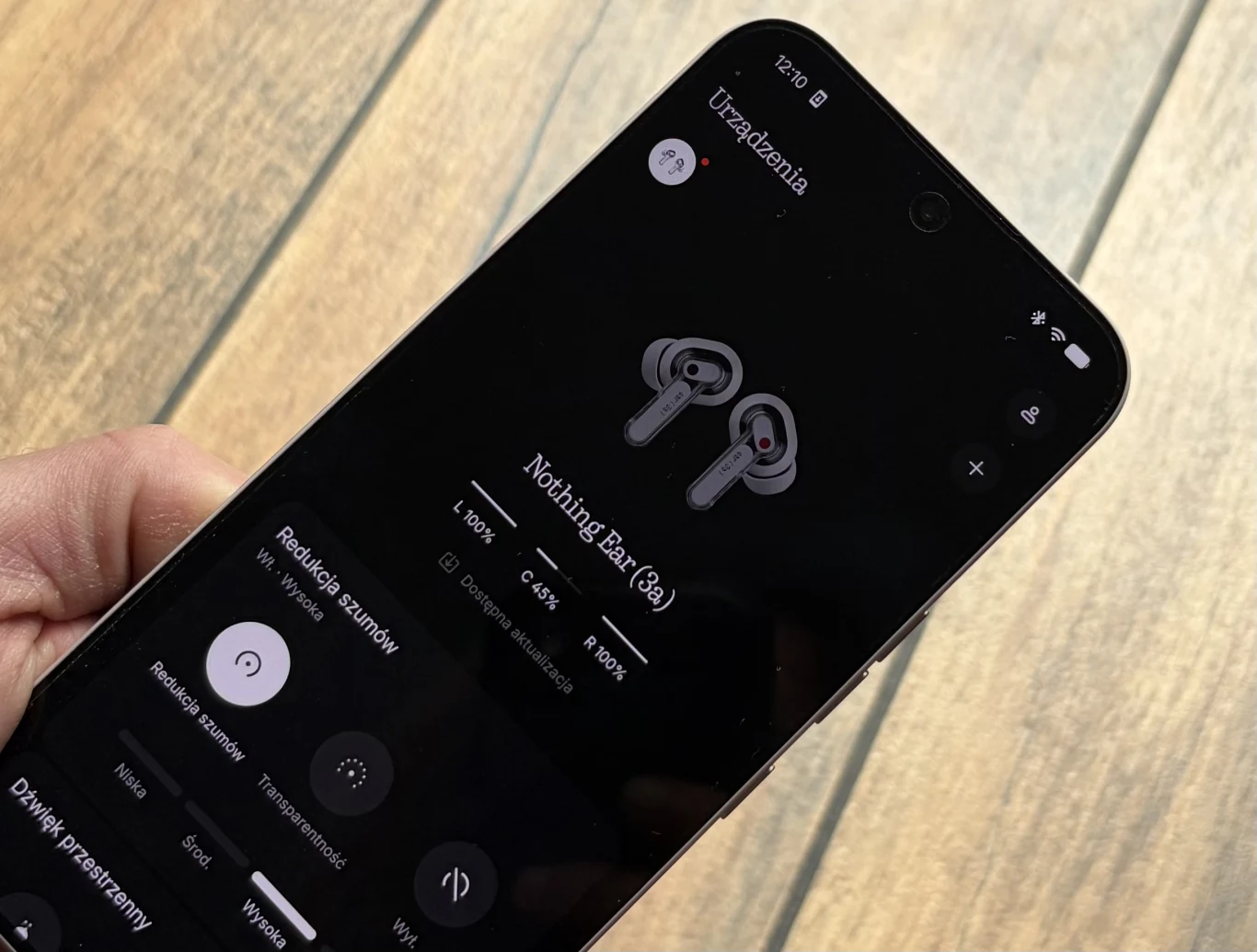 Smartfon trzymany w dłoni z wyświetloną aplikacją do zarządzania słuchawkami Nothing Ear (3), widoczną ikoną słuchawek oraz opcjami regulacji dźwięku i redukcji szumów na tle drewnianej podłogi.