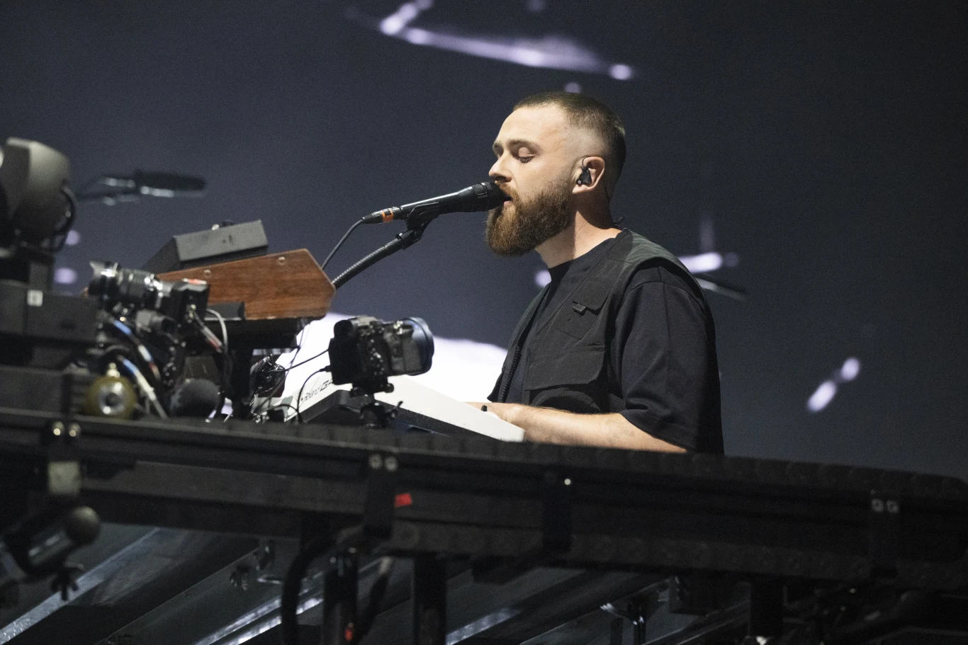 Disclosure Członek Disclosure z brodą śpiewa do mikrofonu, otoczony sprzętem muzycznym na scenie koncertowej.