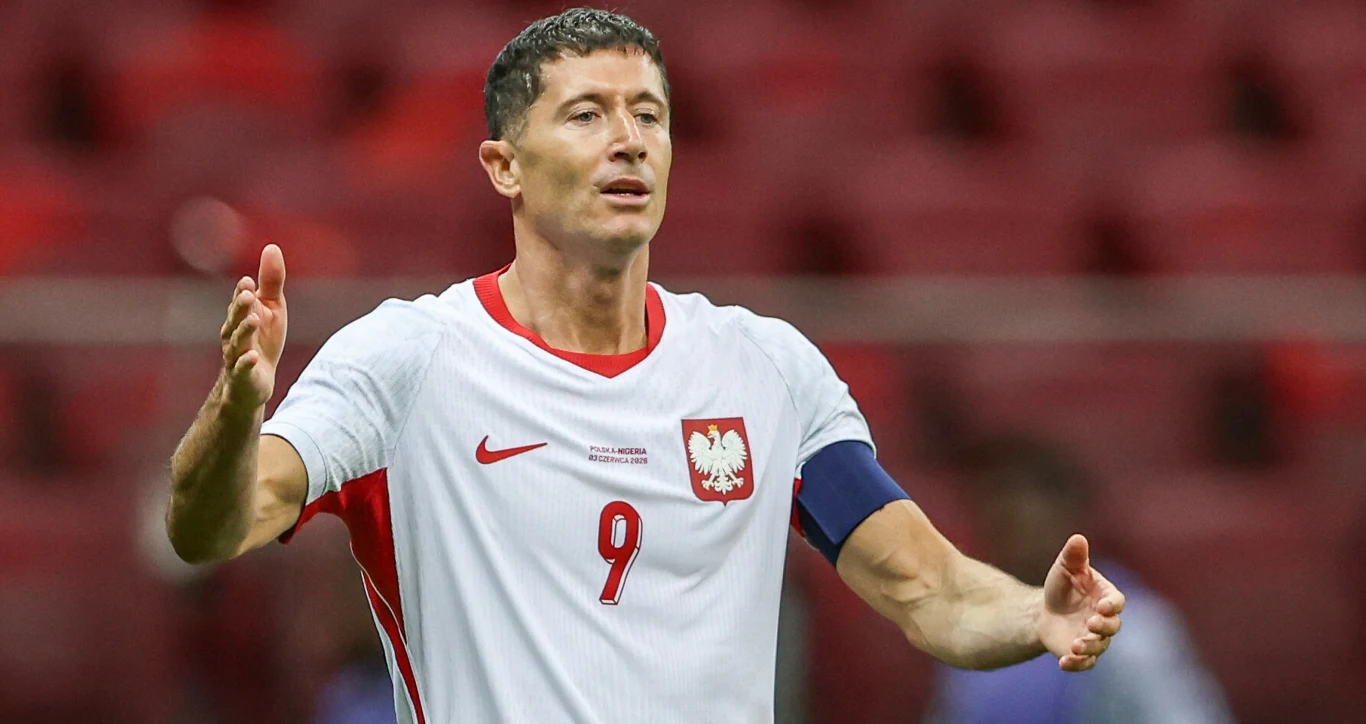 Piłkarz w białej koszulce z numerem 9 i opaską kapitana stoi z szeroko rozłożonymi ramionami na stadionie, prezentując zdeterminowaną postawę.