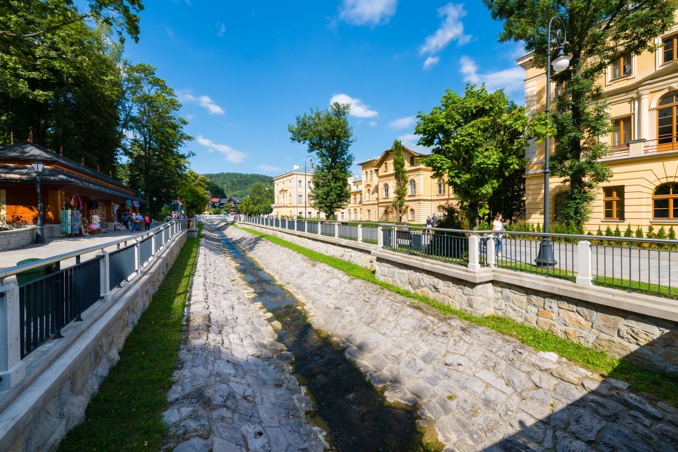 Krynica Zdrój. Kryniczkanka płynąca wzdłuż deptaka w Krynicy Kamienny kanał wodny otoczony żeliwnymi barierkami, po lewej stronie deptak z ludźmi, po prawej stronie zabytkowe budynki wśród zielonych drzew, w tle wzgórza porośnięte lasem pod niebieskim niebem z niewielką ilością chmur.