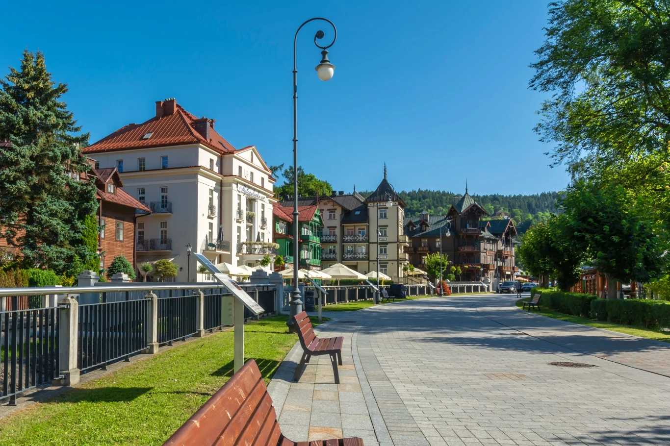 Krynica Zdrój Spacerowy deptak z drewnianymi ławkami, otoczony eleganckimi kamienicami i pensjonatami w górskim kurorcie, w słoneczny, letni dzień.