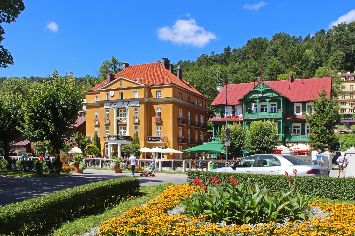 Krynica pełna jest zabytkowych willi Kolorowe kamienice z ogródkami kawiarnianymi, zadbana rabata kwiatowa na pierwszym planie, zaparkowane samochody i otoczenie zielonych drzew.