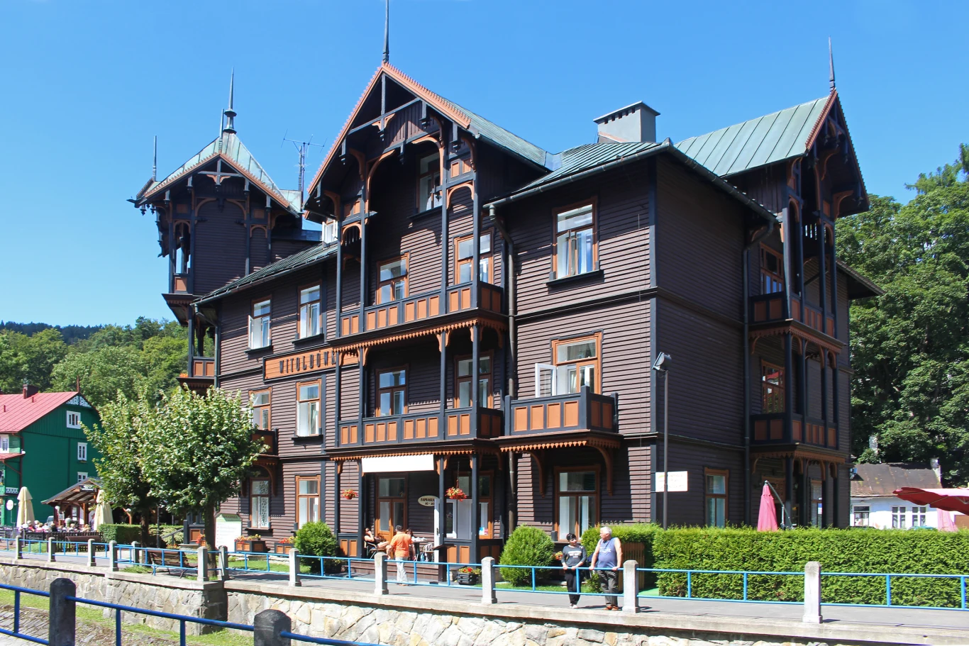 W Krynicy znajduje się wiele ciekawych budynków Drewniany, stylowy budynek z licznymi balkonami i dekoracyjnymi elementami architektonicznymi, otoczony zielenią, z grupką osób spacerujących w pobliżu.