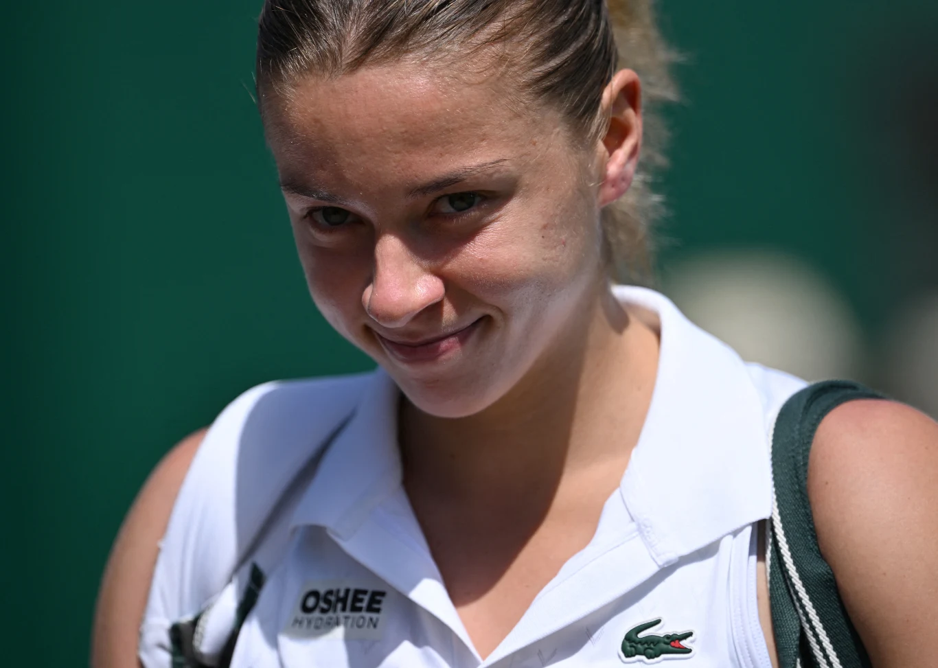 Uśmiechnięta kobieta w sportowej koszulce z logo Lacoste i OSHEE, na tle zielonego tła.