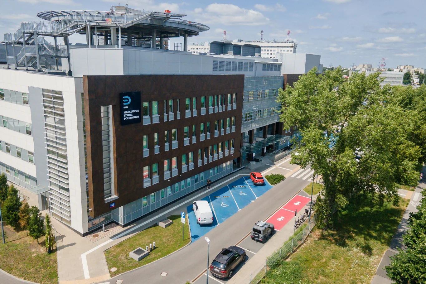 Szpital Południowy Nowoczesny budynek Szpitala Południowego z parkingiem, miejscami dla niepełnosprawnych oraz zielenią.