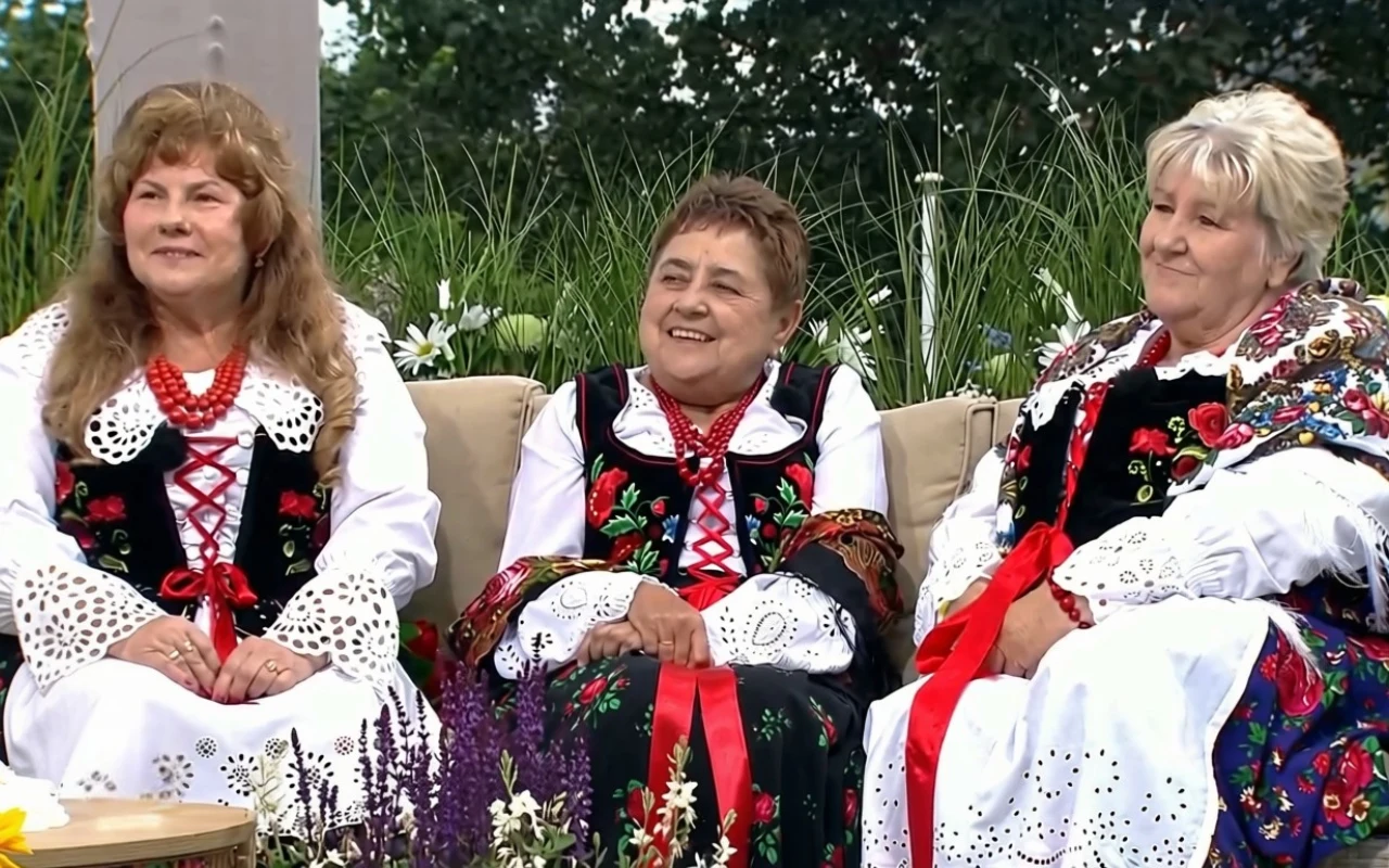 Gościniami wakacyjnego wydania "halo tu polsat" w Zatorze były członkinie Stowarzyszenia Gospodyń Gminy Wieprz Trzy kobiety ubrane w tradycyjne stroje ludowe z kolorowymi haftami i czerwonymi wstążkami siedzą obok siebie na ławce, otoczone kwiatami i zielenią.