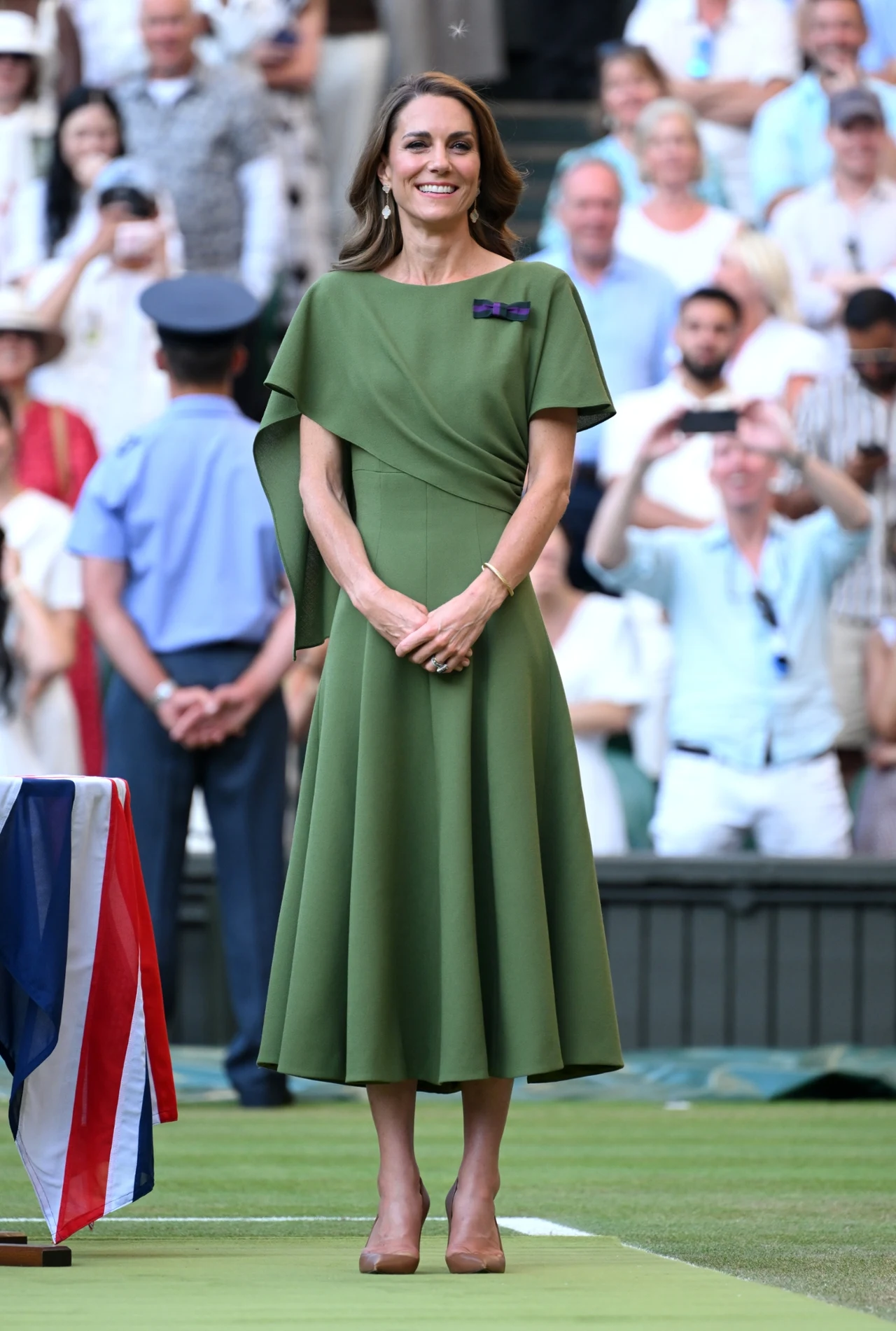 Elegancko ubrana kobieta w zielonej sukience stoi na korcie tenisowym, obok ozdobionego flagą stołu, w otoczeniu tłumu widzów.