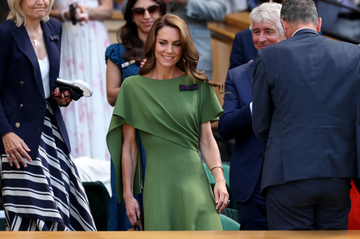 Elegancka kobieta w zielonej sukience z broszką stoi wśród ludzi, uśmiechając się i kierując się do przodu w oficjalnym otoczeniu.