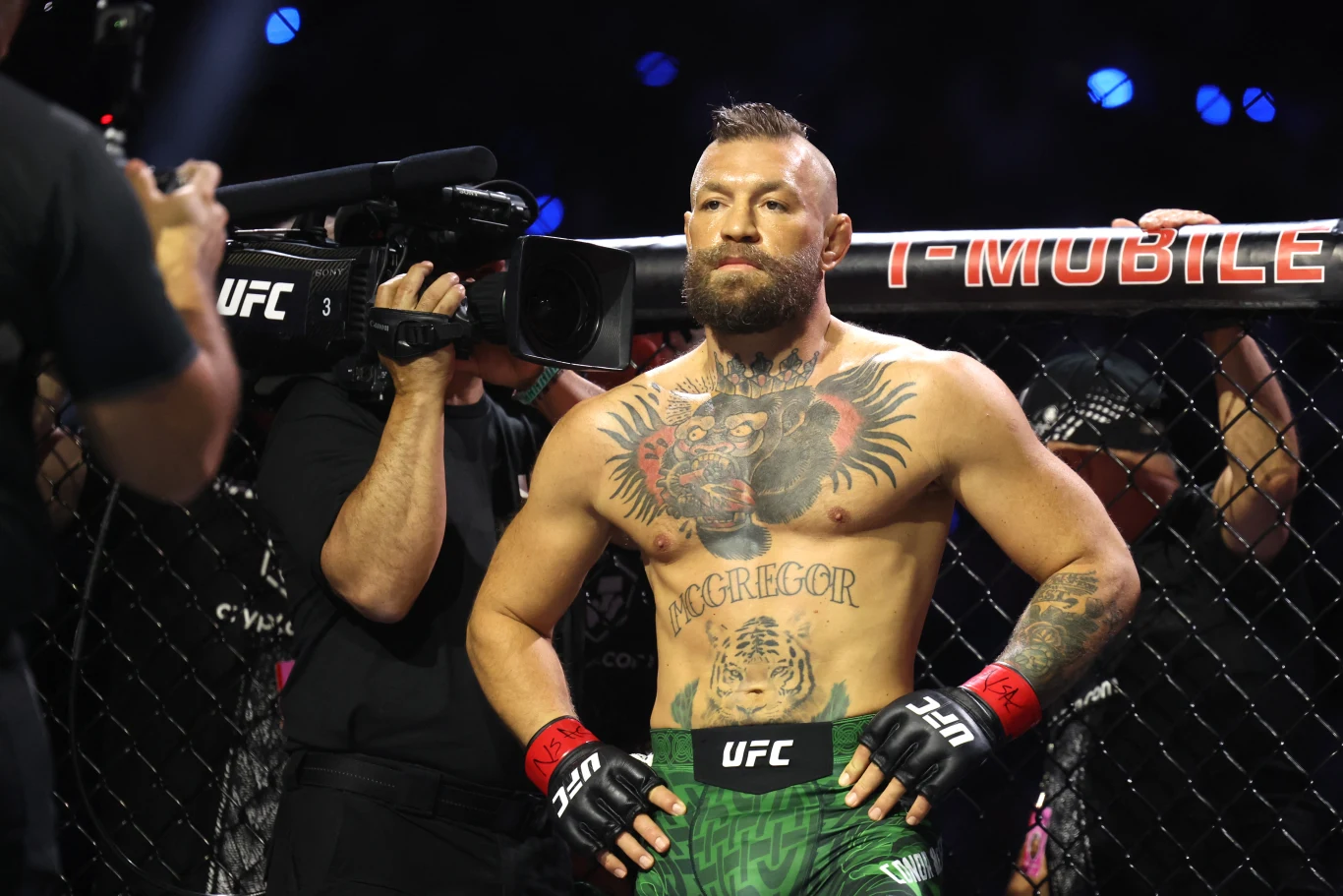 Zawodnik MMA w rękawicach i spodenkach UFC stoi oparty o siatkę oktagonu, otoczony przez kamerzystów i obsługę wydarzenia.