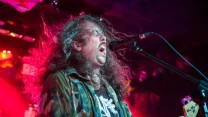Mężczyzna z długimi, rozwianymi włosami w wojskowej kurtce śpiewa do mikrofonu podczas koncertu rockowego, trzymając gitarę basową, a scena oświetlona jest intensywnym, kolorowym światłem.