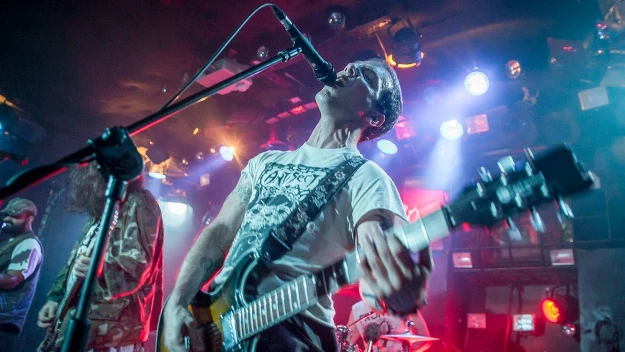 Mężczyzna grający na gitarze elektrycznej i śpiewający do mikrofonu podczas koncertu, w otoczeniu kolorowych świateł scenicznych i innych muzyków.