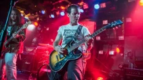 Dwóch muzyków grających na gitarach na tle kolorowych świateł scenicznych, w otoczeniu sprzętu nagłaśniającego i perkusji.