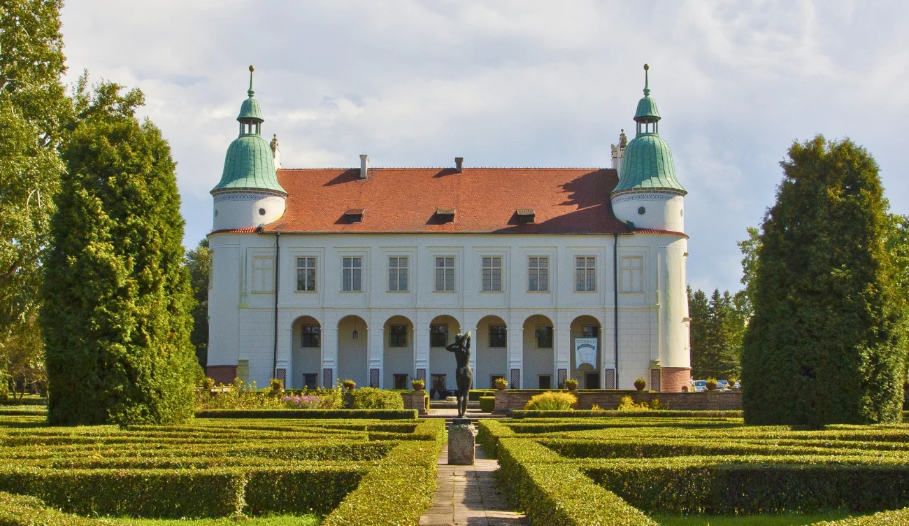 Ile trwa zwiedzanie zamku w Baranowie Sandomierskim? Zamek w Baranowie Sandomierskim, biały budynek z dwiema zielonymi wieżami i czerwonym dachem, otoczony ogrodem.