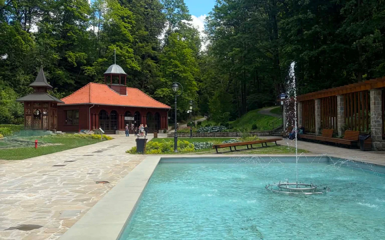 Krynica-Zdrój Zabytkowy budynek z czerwonym dachem otoczony zielenią, fontanna z wodotryskiem w centrum placu oraz drewniana altana i ławki dookoła.