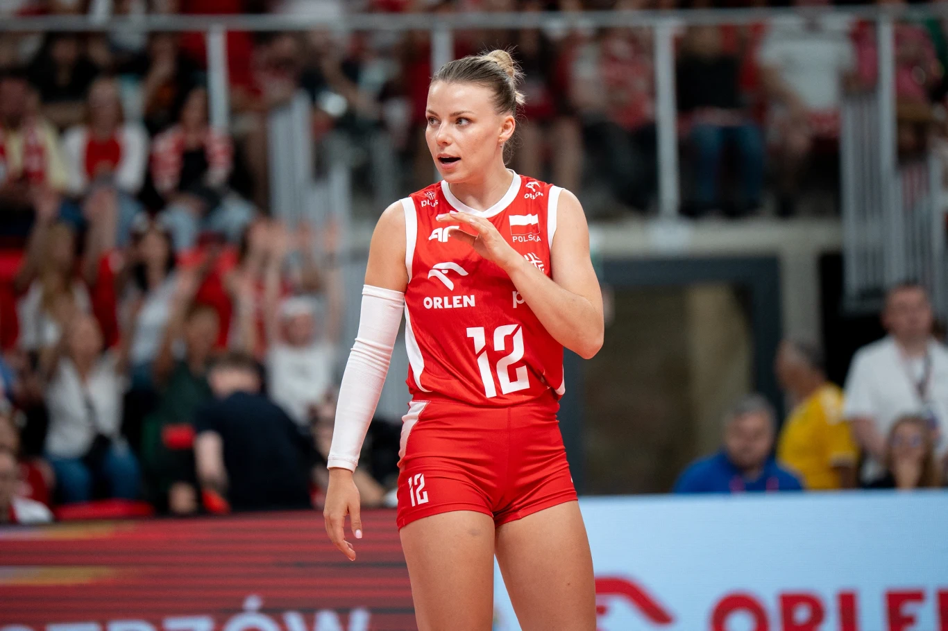 Siatkarka w czerwonym stroju z numerem 12 stoi na boisku, wyrażając emocje i gotowość do gry, w tle widoczni kibice oraz elementy hali sportowej.