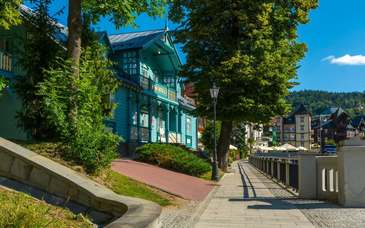 Krynica-Zdrój ma swój klimat Drewniana, niebieska willa w stylu secesyjnym otoczona zielenią przy szerokiej chodnikowej alejce, z widocznymi w tle kolejnymi zabudowaniami uzdrowiskowymi i pagórkowatym krajobrazem.