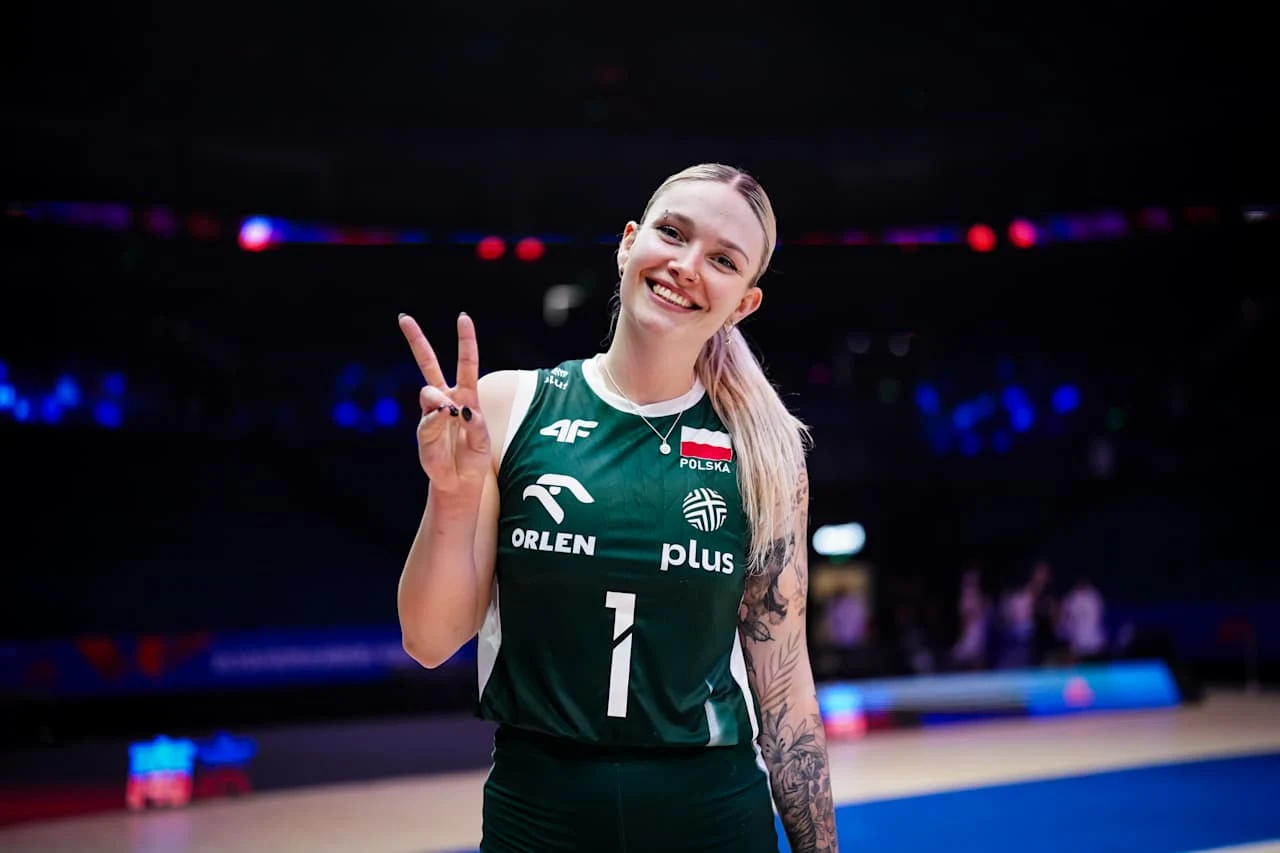 Julia Szczurowska Uśmiechnięta siatkarka w zielonym stroju reprezentacji Polski pokazuje znak pokoju na tle hali sportowej.