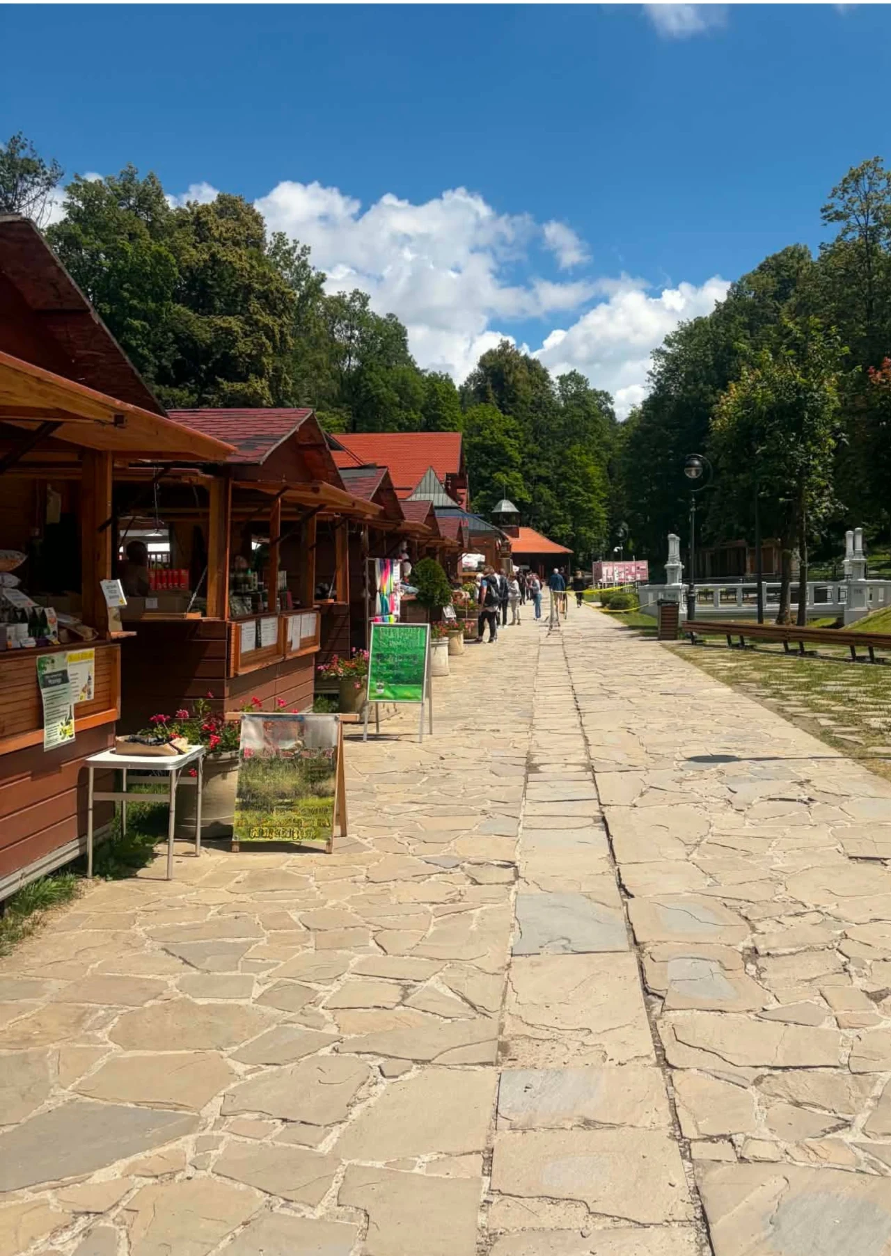 W Krynicy znajdziesz liczne stragany z pamiątkami i regionalnymi wyrobami Drewniane stragany ustawione wzdłuż szerokiej, kamiennej alejki, otoczonej zielenią drzew i jasnym niebem z białymi chmurami.