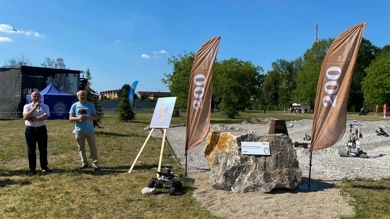 Pole Testowe Łazików Marsjańskich w Józefosławiu. Dwie osoby stoją obok dużego głazu z tabliczką informacyjną, po bokach ustawione są dwie flagi z napisem '200', a na trójnogu znajduje się plansza; w tle teren zielony oraz kilka małych pojazdów terenowych na żwirowej nawierzchni.