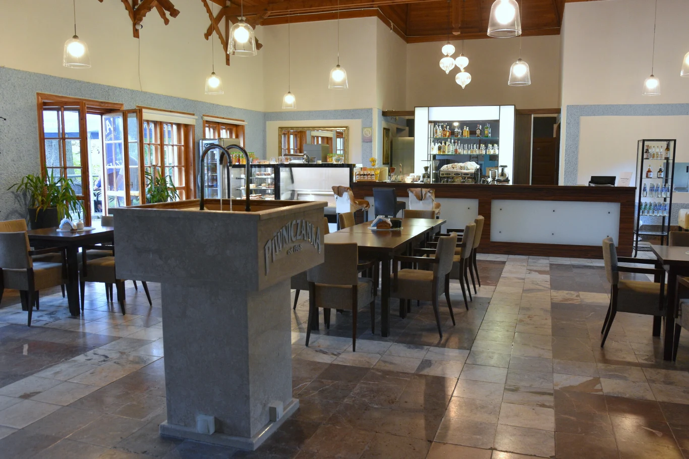 Nowoczesna sala restauracyjna z centralnie ustawionymi stołami i krzesłami, eleganckim barem w głębi, przeszkloną lodówką na napoje oraz dużymi oknami zapewniającymi dostęp naturalnego światła.