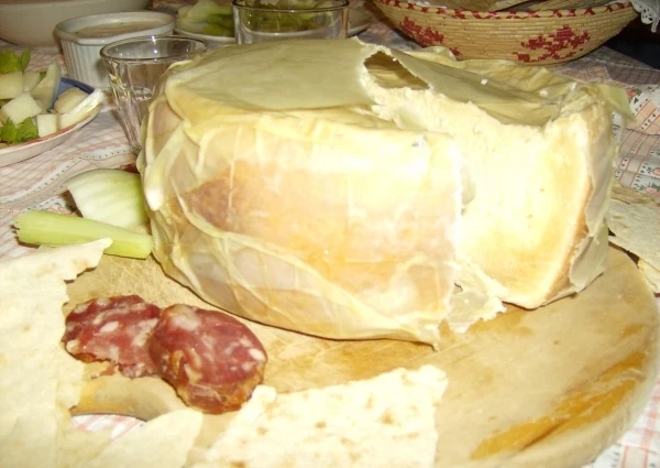 Duży okrągły ser z odkrojonym kawałkiem, obok plasterki kiełbasy i pieczywo na drewnianej desce.