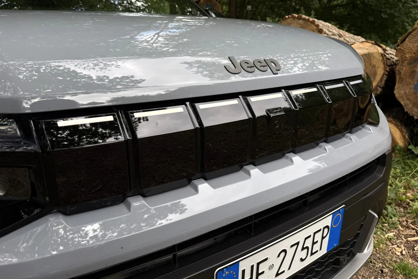 Przedni grill samochodu Jeep z charakterystycznym napisem marki i włoską tablicą rejestracyjną na tle ściętych pni drzew.
