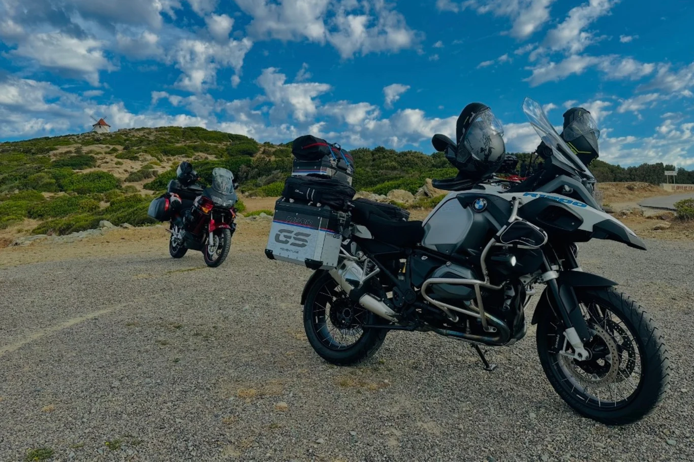 Dwa motory turystyczne BMW GS zaparkowane na kamienistej drodze pośród pagórkowatego, zielonego terenu pod niebem z rozproszonymi chmurami.