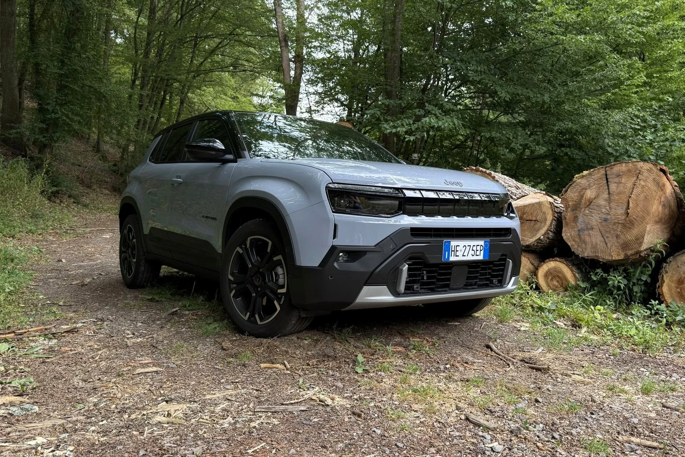 Nowoczesny, szary SUV Jeep zaparkowany na leśnej drodze obok pni świeżo ściętych drzew, otoczony gęstą zielenią lasu.