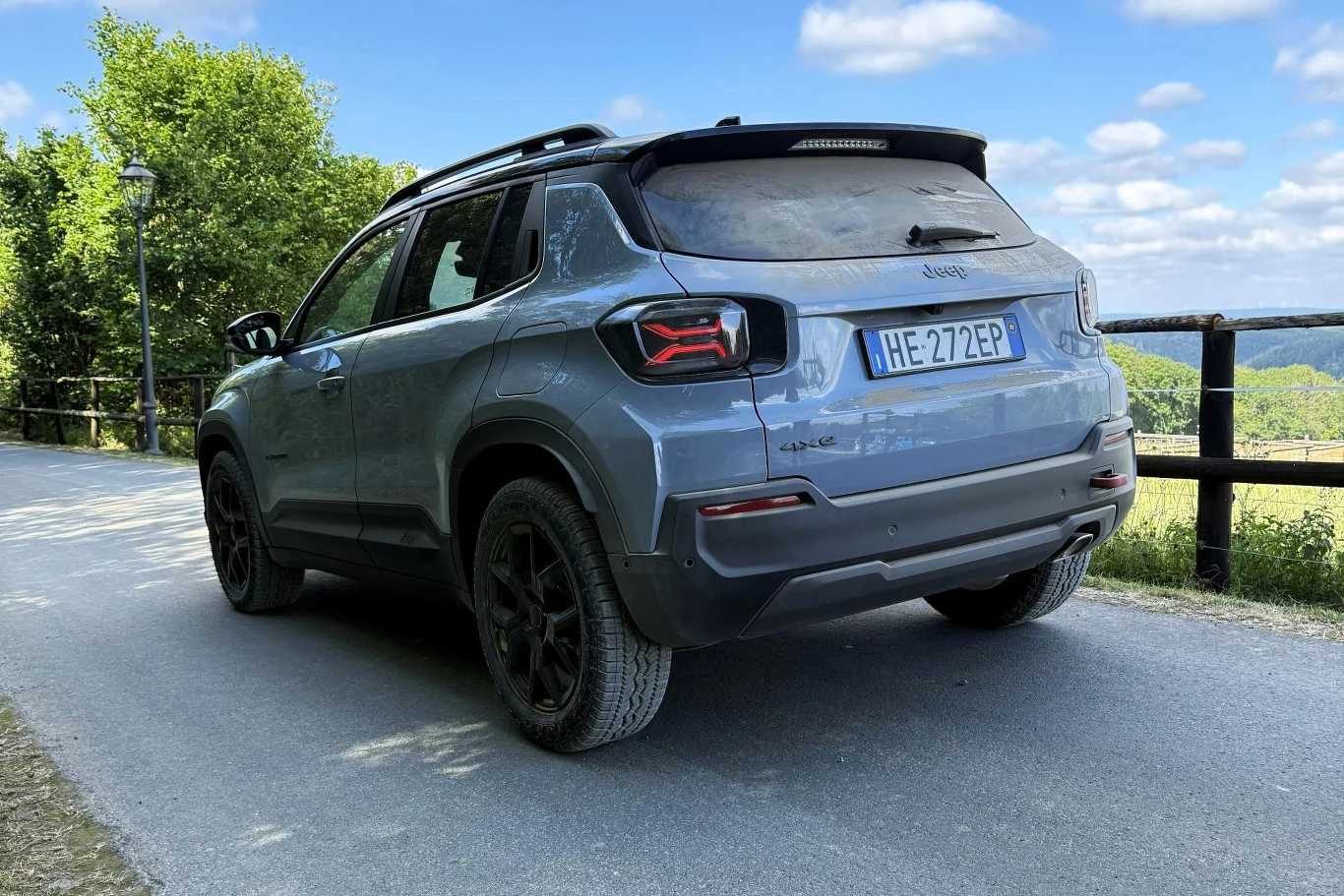 Szary samochód typu SUV marki Jeep zaparkowany na asfaltowej drodze, otoczony barierką i zielenią w słoneczny dzień.