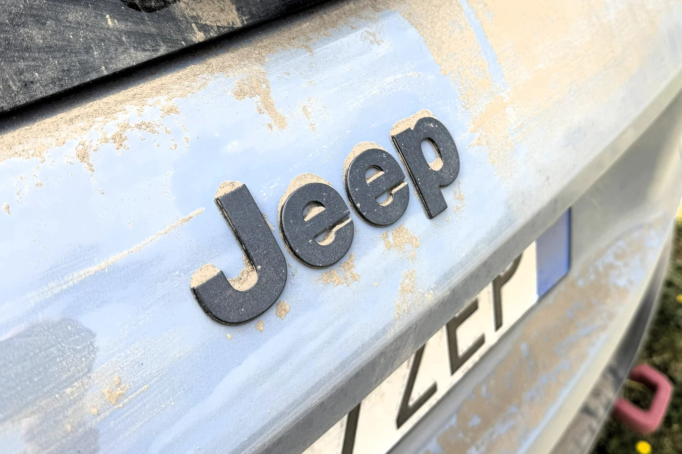 Logo marki Jeep na zakurzonej klapie bagażnika samochodu, widoczny fragment tablicy rejestracyjnej.