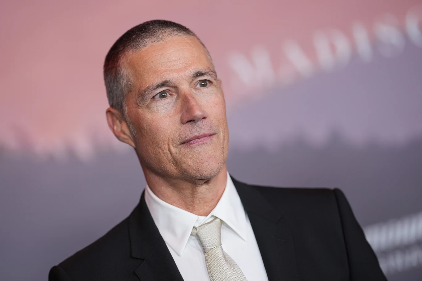Matthew Fox w marcu 2026 roku Mężczyzna w średnim wieku z krótkimi włosami, ubrany w elegancki czarny garnitur, białą koszulę i jasny krawat, stoi na tle rozmazanego, różowego tła.