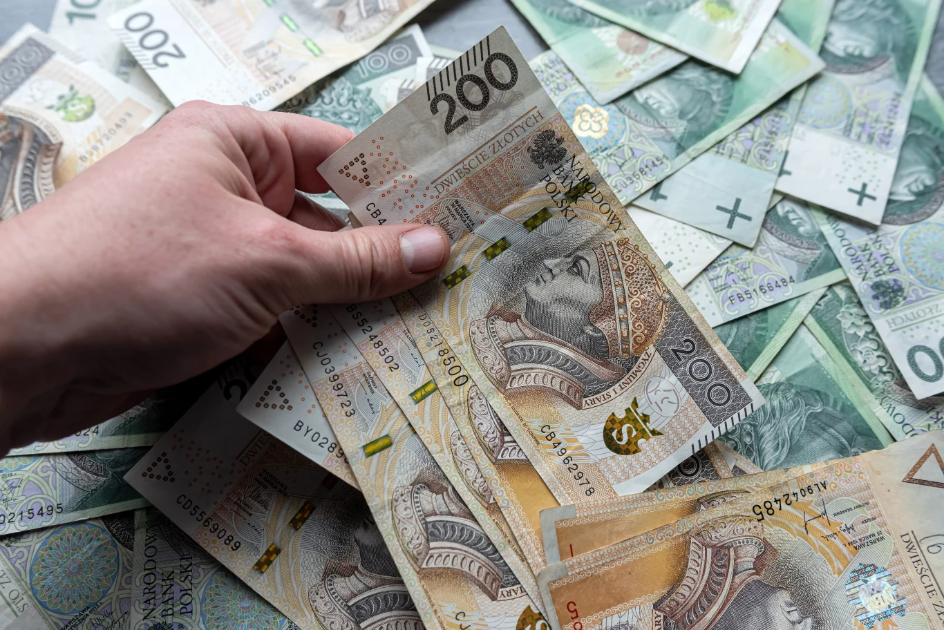 Popularny rytuał na pieniądze może pomóc w odzyskaniu kontroli nad finansami. Jedno działanie może przynieść szczęście. Dłoń trzymająca kilka banknotów 200 zł na tle rozłożonych polskich banknotów o różnych nominałach.