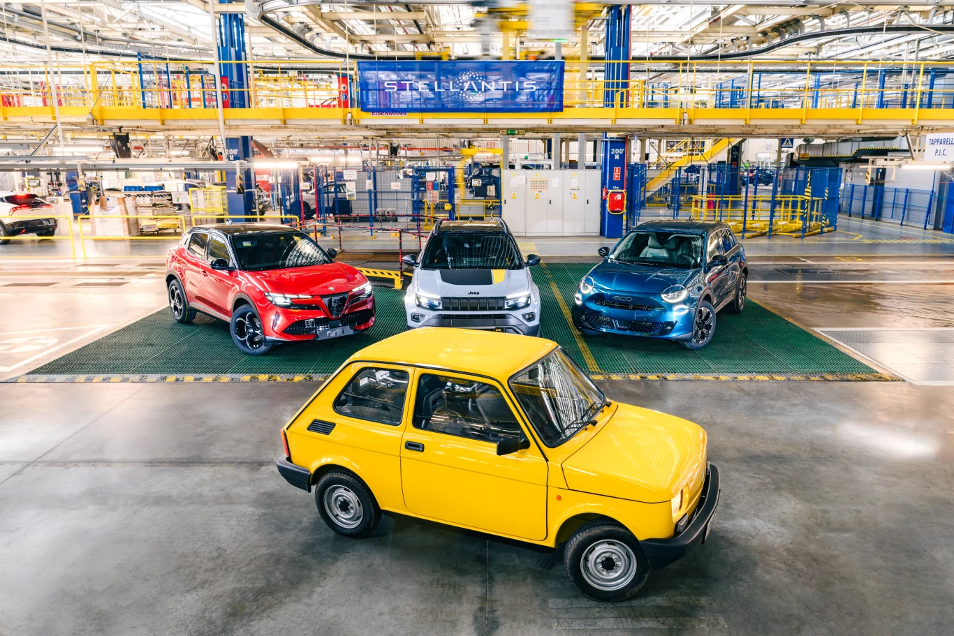 Żółty Fiat 126p wyeksponowany na pierwszym planie hali fabrycznej, w tle nowoczesne modele samochodów ustawione obok siebie na zielonej platformie produkcyjnej.