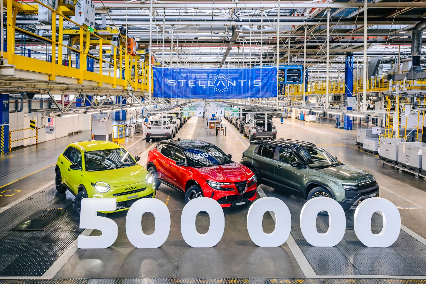 Trzy różnokolorowe samochody ustawione w hali produkcyjnej obok dużego napisu 500 000, w tle logo Stellantis oraz linia montażowa.