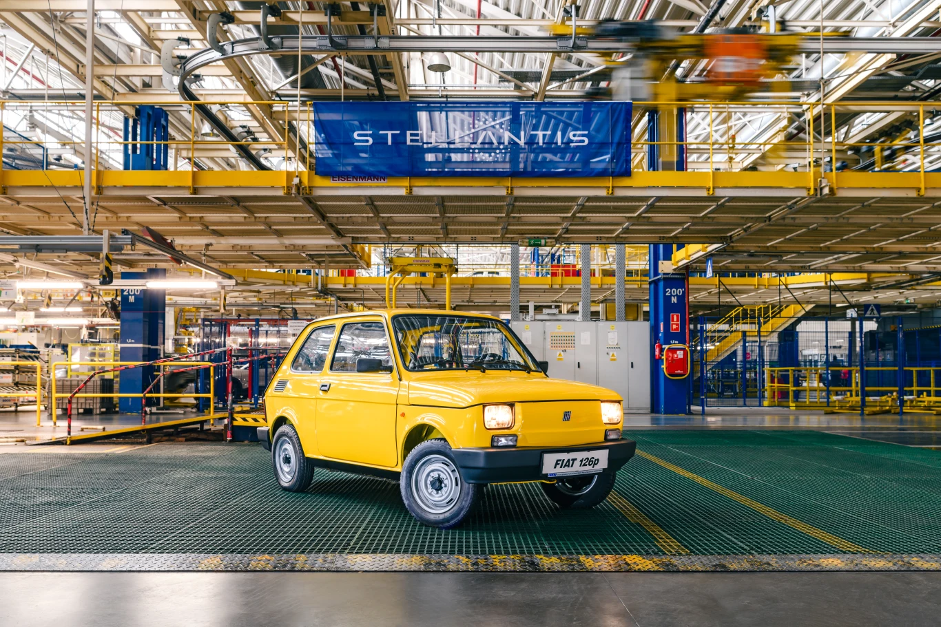 Żółty Fiat 126p ustawiony na tle nowoczesnej hali produkcyjnej z widocznym oznaczeniem Stellantis nad pojazdem.
