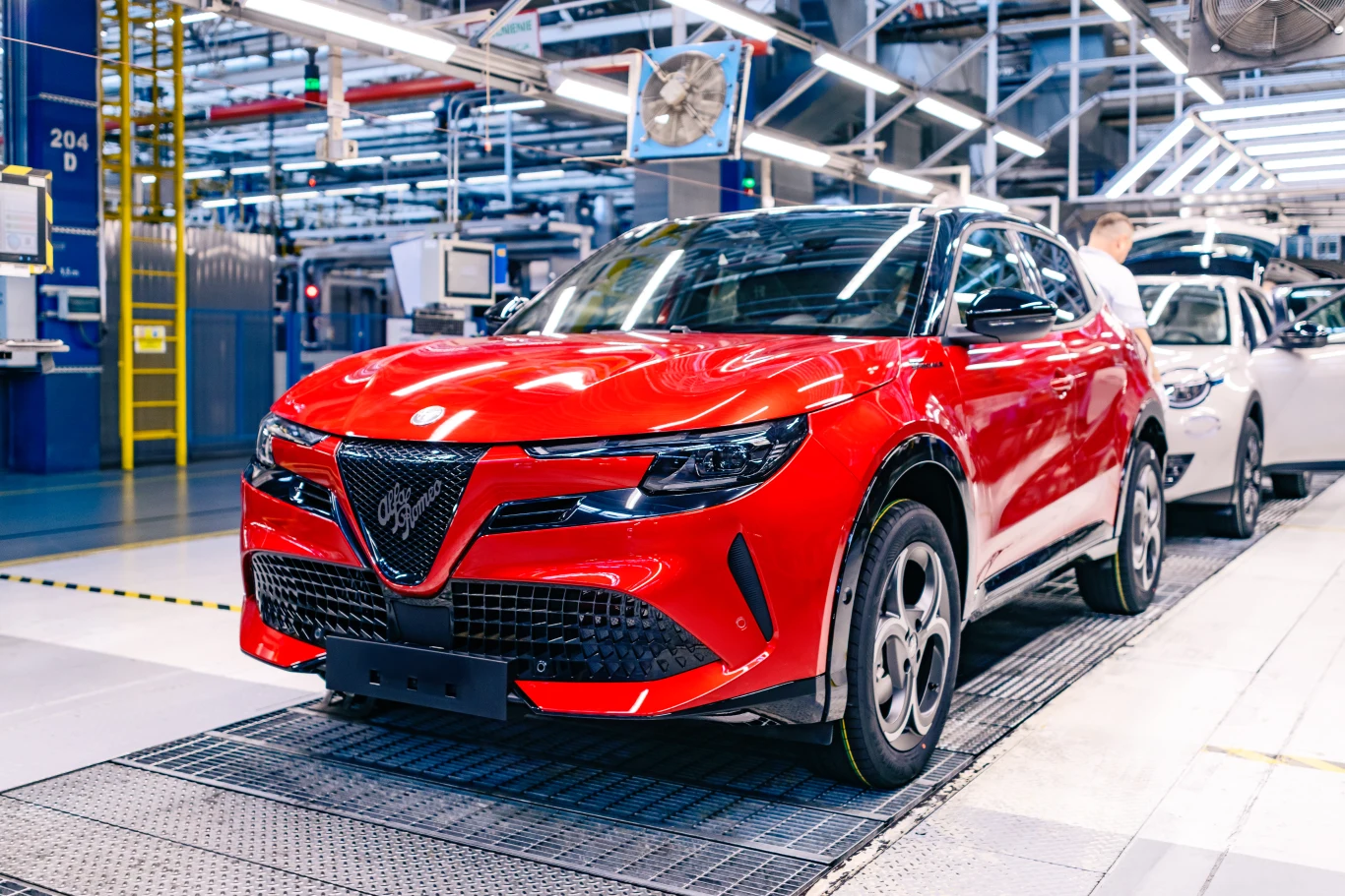 Czerwony samochód Alfa Romeo stojący na linii montażowej nowoczesnej fabryki motoryzacyjnej, w otoczeniu specjalistycznych urządzeń produkcyjnych oraz innego pojazdu w tle.