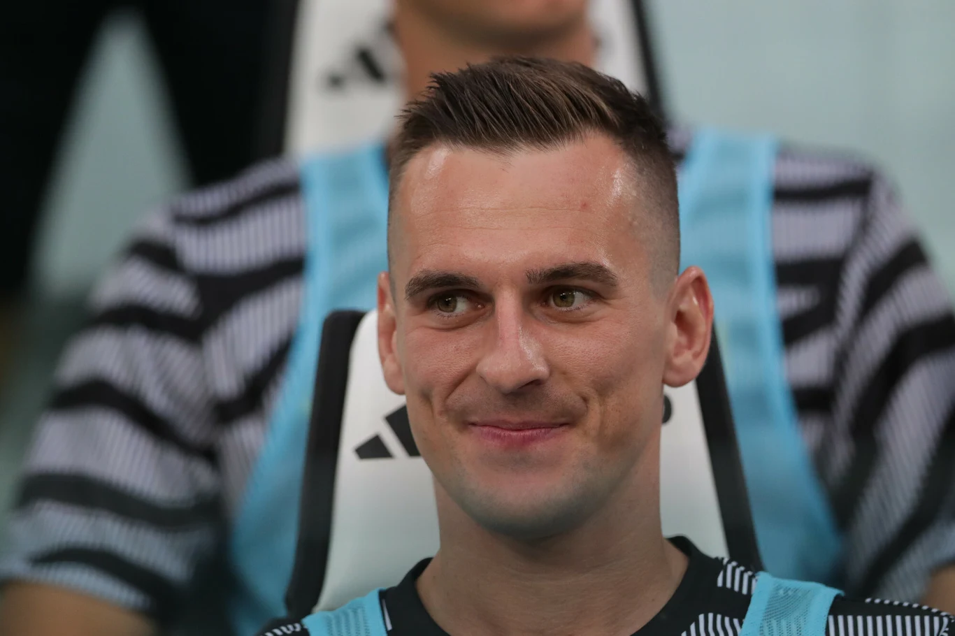 Mężczyzna w sportowym stroju z uśmiechem na twarzy, siedzący na ławce rezerwowych podczas meczu piłkarskiego.