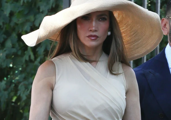 Jennifer Lopez Jennifer Lopez w eleganckiej, kremowej sukni bez rękawów i dużym, szerokim kapeluszu w odcieniu beżu, stojąca na zewnątrz w świetle dziennym.