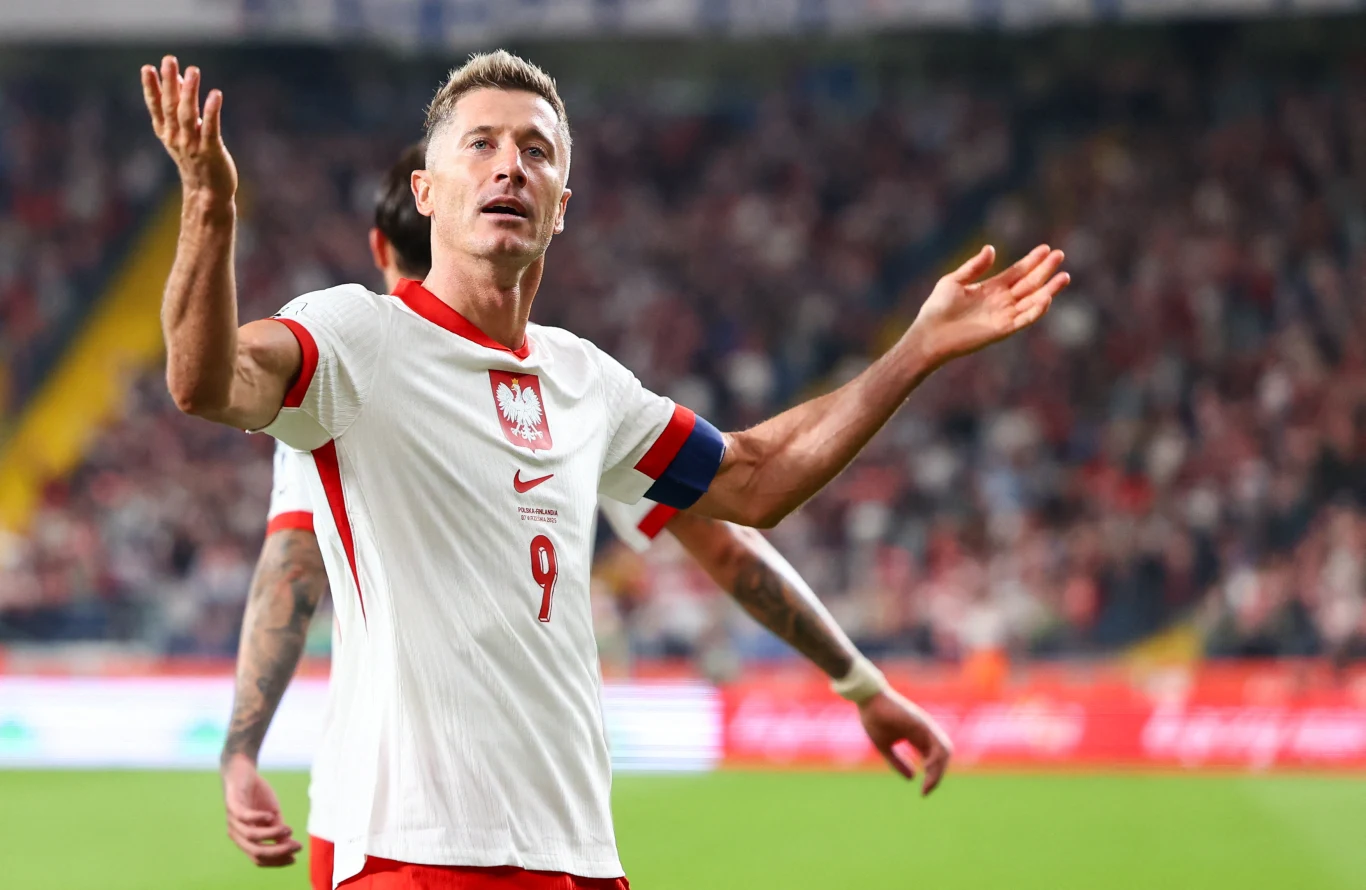 Piłkarz w białej koszulce z numerem 9 i opaską kapitana gestykuluje z uniesionymi rękami na stadionie w trakcie meczu.