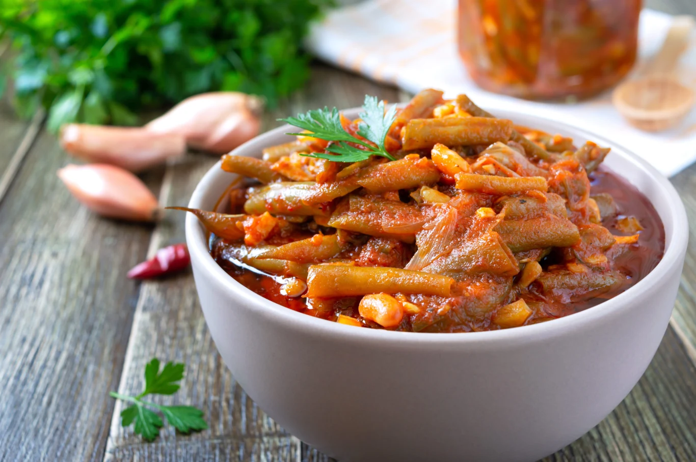 Fasolka po gruzińsku to warzywa zatopione w ostrym, aromatycznym sosie. Porcja fasolki szparagowej po gruzińsku w sosie pomidorowym z natką pietruszki, w misce na drewnianym stole.