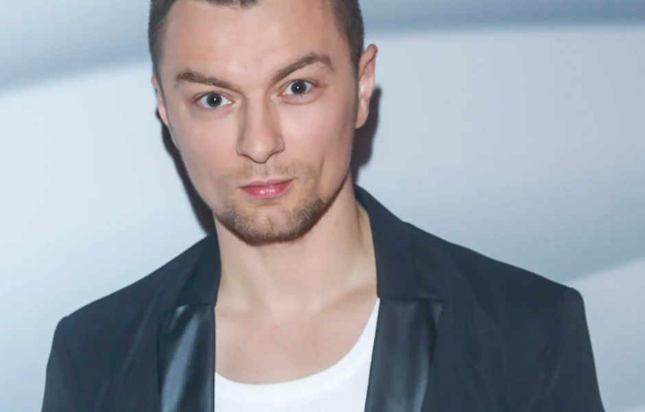Liber Młody mężczyzna z zarostem, ubrany w biały t-shirt i ciemną marynarkę, patrzy bezpośrednio w obiektyw z lekko zaskoczonym wyrazem twarzy.